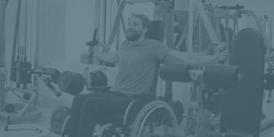 En mand i kørestol er ved at udføre en træningsøvelse, hvor arme og brystmuskulatur trænes.