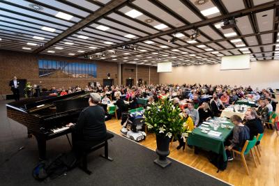 Billedet viser en fyldt plenumsal til Handicaprådenes Dag, hvor godt 200 deltagere sidder til bords. I billedets forgrund sidder en blind pianist og spiller flygel.