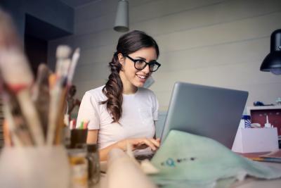 Ung kvinde med briller arbejder ved computer. I forgrunden står et glas med pensler.
